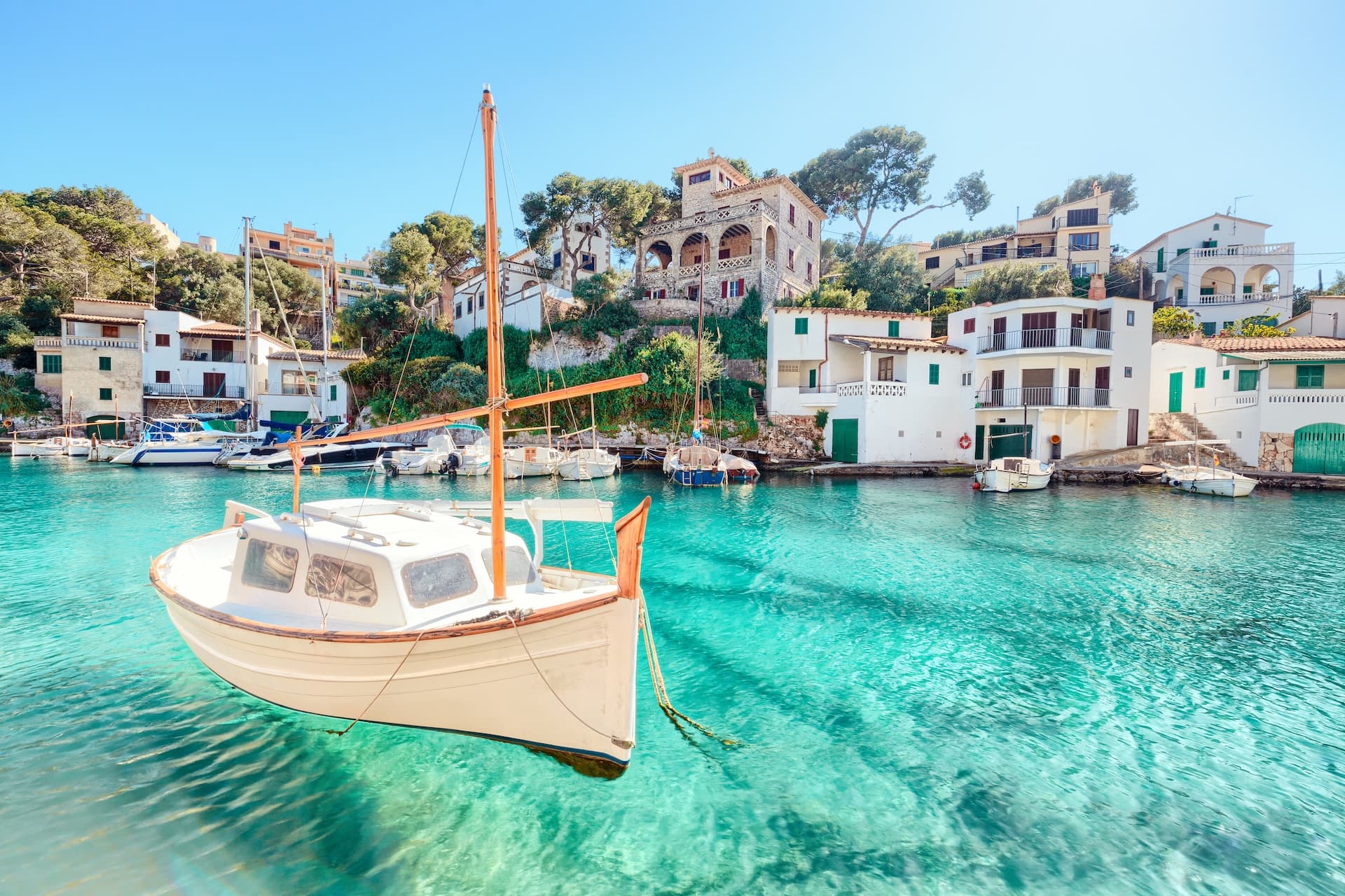 Small white boat floating in turquoise water near white buildings in Cala Figuera, Mallorca.